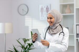 giovane bellissimo femmina medico nel hijab utilizzando tavoletta computer, medico lavoratore nel bianca medico cappotto e stetoscopio dentro clinica ufficio a posto di lavoro, sorridente musulmano donna utilizzando in linea applicazione. foto