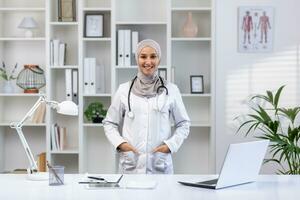 ritratto di contento arabo femmina medico nel hijab in piedi a tavolo nel ospedale ufficio con stetoscopio in giro collo, mani nel tasche, sorridente e guardare a telecamera. foto