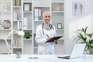 ritratto di un' musulmano femmina medico nel un' hijab in piedi nel un' moderno ufficio, Tenere un' tavoletta con documenti nel sua mani, sorridente e guardare a il telecamera. foto