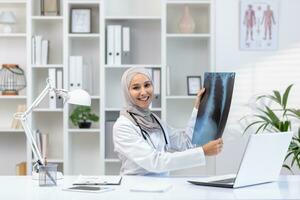 arabo musulmano donna medico seduta a tavolo nel ufficio nel privato clinica Tenere raggi X nel mani conduzione consultazione sorridente guardare a telecamera. foto