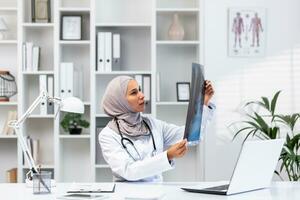 giovane riuscito femmina medico nel hijab Lavorando nel clinica dentro medico ufficio, musulmano donna è arrabbiato e pensiero raggi X immagine seduta a posto di lavoro con il computer portatile. foto