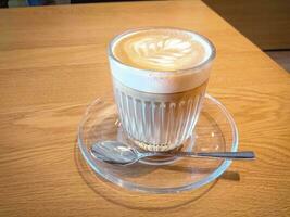 caldo cremoso cappuccino caffè con schiuma servito nel un' costine bicchiere su un' bicchiere piatto, caldo latte macchiato caffè bevanda nel bicchiere con crema latte su di legno tavolo, prima colazione bevanda mattina concetto. foto