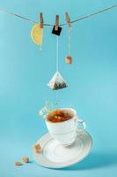 bustina di tè, limone e zucchero appesi alla corda su spruzzi di tè su sfondo blu. concetto di natura morta creativa.