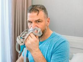 infelice uomo scioccato con problemi respiratori cronici sorpreso dall'uso della macchina cpap seduto sul letto in camera da letto. assistenza sanitaria, cpap, terapia dell'apnea ostruttiva del sonno, concetto di russamento foto