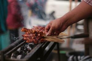 pollo satay al di sopra di tradizionale carbone fuoco. il satay ustioni con Fumo e un appetitoso aspetto esteriore. mani cucinando satay su il griglia. persone siamo già fare la fila. foto