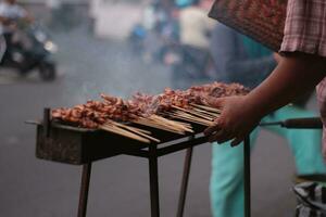 pollo satay al di sopra di tradizionale carbone fuoco. il satay ustioni con Fumo e un appetitoso aspetto esteriore. mani cucinando satay su il griglia. persone siamo già fare la fila. foto