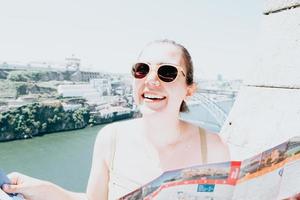 Giovane donna turista con occhiali da sole alla ricerca di una mappa nel mezzo di una strada e sorridente pensando alla fotocamera, giornata estiva, soleggiata, porto, città mediterranea, foto