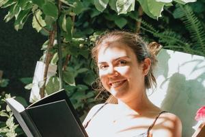 donna con in mano un libro che sorride alla telecamera mentre è sdraiata su una sedia durante una giornata di sole, copia spazio, relax e concetti di hobby foto