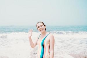 giovane donna in costume da bagno saluta la telecamera durante una giornata di sole in spiaggia con l'oceano come sfondo, libertà e concetto di vacanza, spazio di copia foto