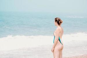 giovane donna in costume da bagno di fronte all'oceano che si gira verso la macchina fotografica e sorride, copia spazio, libertà e concetto futuro