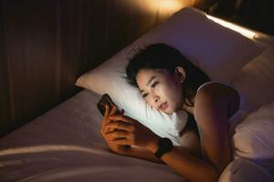 asiatico donna utilizzando mobile Telefono smartphone posa su il letto nel il Camera da letto. assonnato esausto, può non dormire. insonnia, dipendenza concetto. donne scorrimento sociale reti su mobile buio Camera da letto. foto