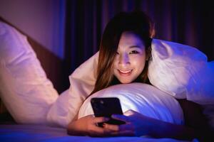 asiatico donna utilizzando mobile Telefono smartphone posa su il letto nel il Camera da letto. donne scorrimento sociale reti su mobile. soddisfare femmina dire bugie nel letto sotto coperta e Guardando film su smartphone. foto