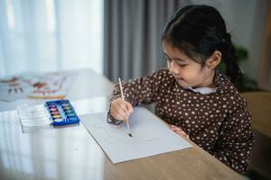 carino poco asiatico bambino sorridente pittura con colorato vernici utilizzando acquerello. asiatico ragazza utilizzando pennello disegno colore. bambino attività stile di vita concetto. foto