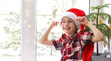 ragazza nel un' Santa cappello si siede su il davanzale di un' Casa vicino il Natale albero e mette caramella canna per sua occhi piace occhiali. bambino è avendo divertimento e fabbricazione facce, in attesa per Natale e nuovo anno foto