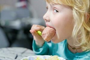 un' poco ragazza ha prima colazione a casa spaghetti con salsicce. poco bionda ragazza mangiare cena con forchetta a tavolo foto