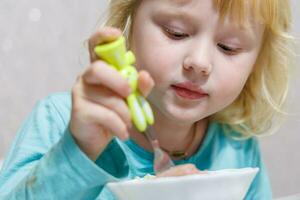 un' poco ragazza ha prima colazione a casa spaghetti con salsicce. poco bionda ragazza mangiare cena con forchetta a tavolo foto