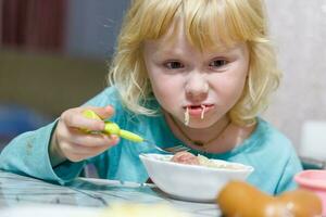 un' poco ragazza ha prima colazione a casa spaghetti con salsicce. poco bionda ragazza mangiare cena con forchetta a tavolo foto