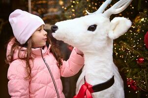 bellissimo ragazzo ragazza ammirazione un' bianca cervo giocattolo, in piedi di Natale albero, illuminato di ghirlande e Natale luci foto