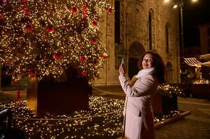 contento donna viaggiatore Fotografare un' Natale albero nel il città quadrato, passeggiando il strada a notte mentre un' luna park foto