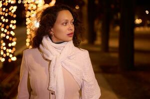 Multi etnico bellissimo giovane donna nel caldo beige cappotto, a piedi a notte nel il strada con Natale luci su fondale foto