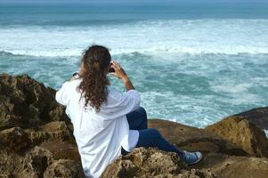 Multi etnico bella donna assunzione foto su sua inteligente mobile Telefono, seduta su il roccia su il atlantico oceano spiaggia