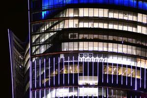 frammento di il bicchiere facciata di un' moderno aziendale edificio a notte. moderno bicchiere ufficio nel città. grande raggiante finestre nel moderno ufficio edifici a notte, nel righe di finestre leggero brilla. foto