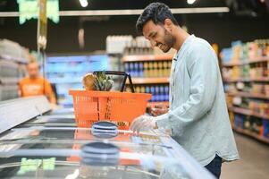 ritratto di contento indiano uomo in piedi nel davanti di il Prodotto contatore nel un' drogheria negozio. uomo acquisto drogheria per casa nel supermercato. foto