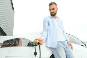 uomo Tenere energia ricarica cavo per elettrico auto nel all'aperto auto parco. e lui S andando per Collegare il auto per il ricarica stazione nel il parcheggio lotto vicino il shopping centro. foto