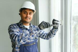 indiano operaio nel tuta da lavoro installazione o regolazione plastica finestre nel il vivente camera a casa foto