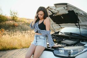 il giovane ragazza rotto il auto e lei ha aperto il cappuccio e provato per riparazione il auto su il strada. un' donna chiamate su un' smartphone con un' auto evacuazione servizio foto