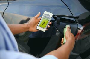 uomo Tenere smartphone mentre ricarica auto a elettrico veicolo ricarica stazione, avvicinamento. foto