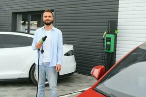 giovane bello uomo Tenere ricarica cavo a elettrico ricarica stazione punto in piedi vicino il suo nuovo auto foto