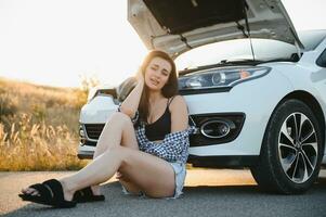 un' giovane ragazza si siede vicino un' rotto auto su il strada con un Aperto cappuccio. foto