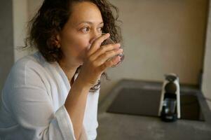 fiducioso Multi etnico donna vestito nel bianca veste, potabile acqua mentre assunzione medicinale, in piedi di cucina contatore foto