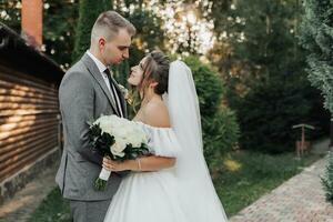 ritratto di il sposa e sposo nel natura. il sposo e il brunetta sposa nel un' bianca lungo vestito siamo in piedi, baci contro il sfondo di conifere. elegante sposo. bellissimo capelli e trucco foto