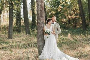 nozze foto nel natura. il sposa e sposo siamo in piedi vicino un' albero sorridente e guardare lontano. il sposo abbracci il suo Amati a partire dal dietro, il sposa detiene un' mazzo. ritratto. estate nozze