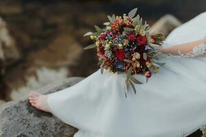 sposa su il banca di il fiume, con spoglio gambe seduta su un' roccia Tenere un' mazzo di vario fiori, rosso Rose e blu fiori e un' lungo bianca vestito foto