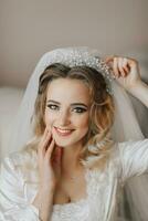 un' bionda sposa è indossare un' raso accappatoio e seduta nel sua camera, in posa e mostrando via sua tiara e velo. bellissimo capelli e trucco, Aperto rompersi. nozze ritratto. sincero Sorridi foto