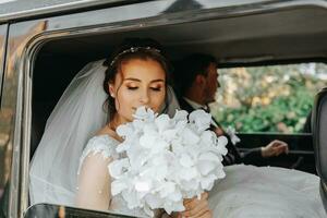 il sposa si siede nel un' nero auto su il nozze giorno con un' mazzo. ritratto di il sposa. lussureggiante bianca pizzo vestito foto