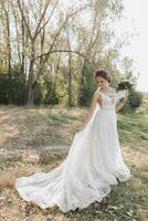 il sposa nel un' bianca vestito con un' lungo treno è a piedi nel il foresta, guardare giù e Tenere un' vestire. il vento colpi il ragazze capelli. il ombre. estate nozze. foto