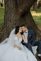 contento giovane coppia, sposa con lungo Riccio capelli nel bianca vestito con lungo treno vicino albero. bellissimo ragazza nel il parco. bellissimo colore. nozze foto sparare