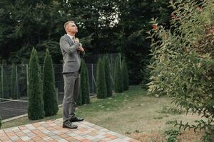 un' uomo nel un' grigio completo da uomo pose nel natura, regola il suo legare. lato Visualizza. un' elegante guarda. Uomini stile. moda. attività commerciale. foto