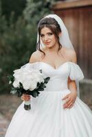 il sposa nel un' voluminoso bianca vestito con Aperto le spalle e un' lungo velo, detiene un' mazzo di bianca Rose e sembra per il lato. ritratto di il sposa. bellissimo trucco. nozze nel natura foto