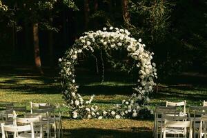 nozze cerimonia nel il foresta. il strada per il il giro arco con fogliame, verdi, verde, e fiori. rustico arredamento. di legno sedie nel il Giardino dietro la casa banchetto la zona. posti a sedere per ospiti. foto