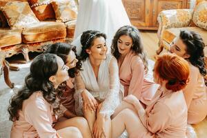 il damigelle d'onore Guarda a il sorridente sposa. il sposa e sua divertimento amici siamo festeggiare un' addio al nubilato festa su il pavimento vicino il nozze vestito nel accoppiamento vestiti. sposa e amici nel il camera foto