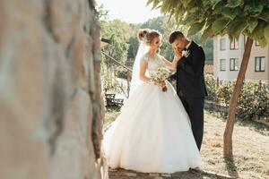 nozze ritratto. il sposo nel un' nero completo da uomo e il bionda sposa In piedi di un' pietra parete sotto un' albero, il sposo baci il della sposa mano. foto sessione nel natura. bellissimo capelli e trucco