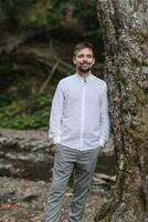 un' giovane e elegante uomo nel un' bianca camicia sta nel il primavera foresta contro il sfondo di un' grande albero, mani nel il suo tasche foto