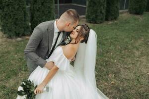 ritratto di il sposa e sposo nel natura. il sposo e il brunetta sposa nel un' bianca lungo vestito siamo in piedi, baci contro il sfondo di conifere. elegante sposo. bellissimo capelli e trucco foto