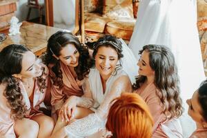 il damigelle d'onore Guarda a il sorridente sposa. il sposa e sua divertimento amici siamo festeggiare un' addio al nubilato festa su il pavimento vicino il nozze vestito nel accoppiamento vestiti. sposa e amici nel il camera foto