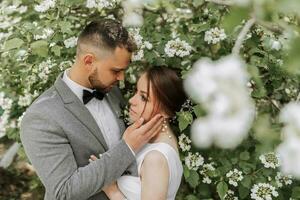 il sposa nel un' bianca sera vestito pose con il sposo con un' bellissimo acconciatura, il sposo delicatamente impacchi il sposa e Leans contro sua contro il sfondo di primavera fiori. foto